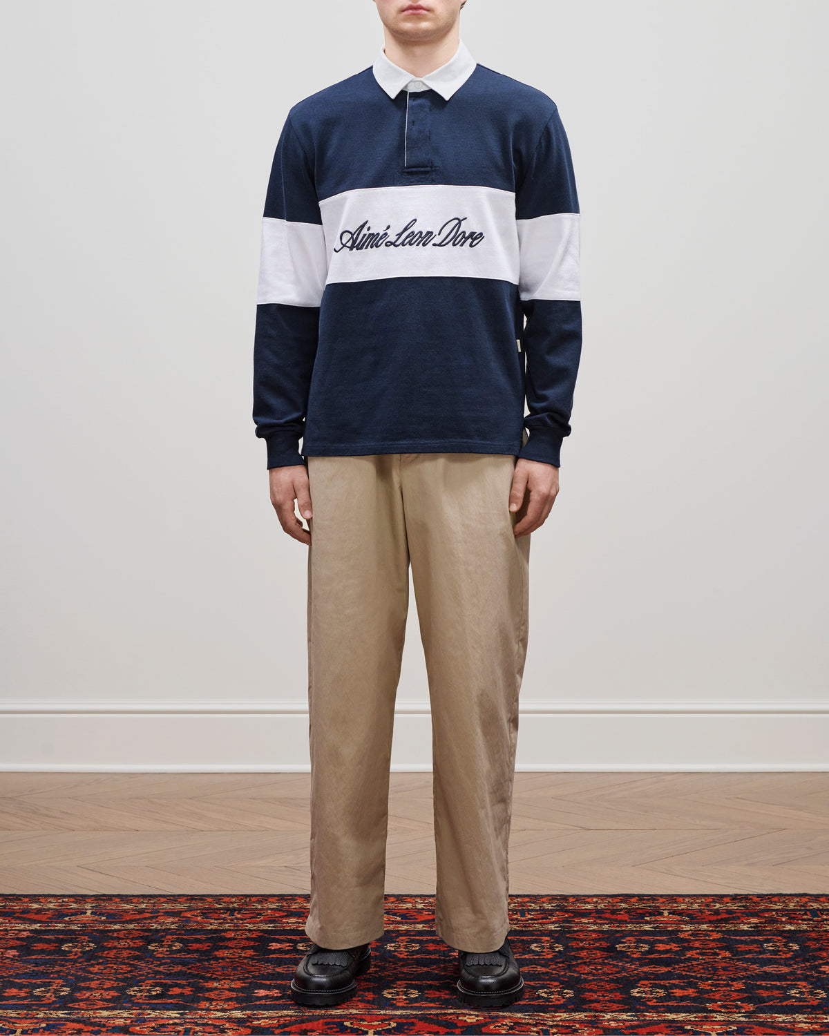 Person wearing a Script Paneled Rugby in navy and white with beige pants, standing on a rug against a plain wall.