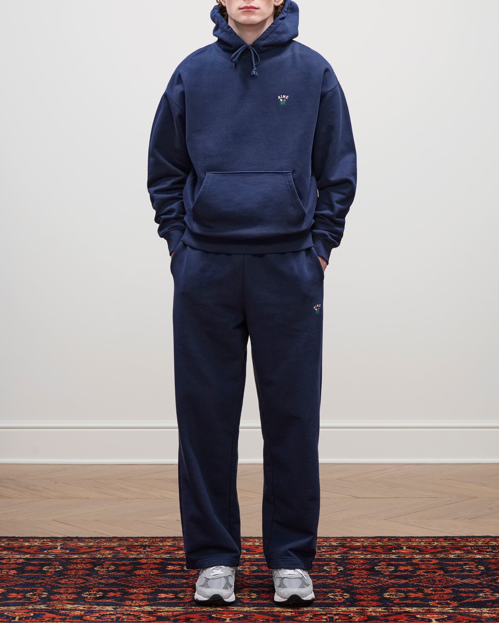 A person wearing a navy Crest Hoodie, sweatpants, and gray sneakers stands on a patterned rug.