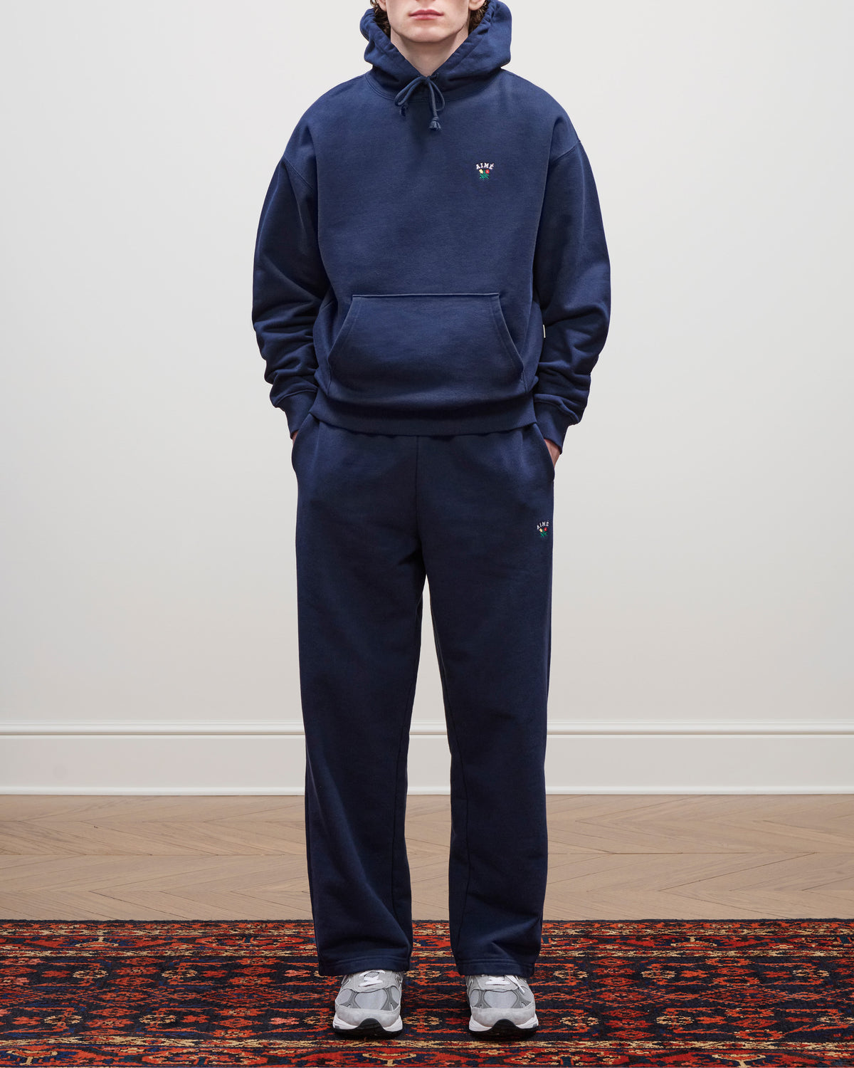 Person wearing Crest Sweatpants and a navy hoodie stands on a patterned rug, paired with gray sneakers.
