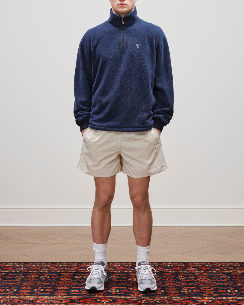 Person in a navy Crest Quarter-Zip Fleece Pullover, beige shorts, white socks, and gray sneakers stands on a rug indoors.