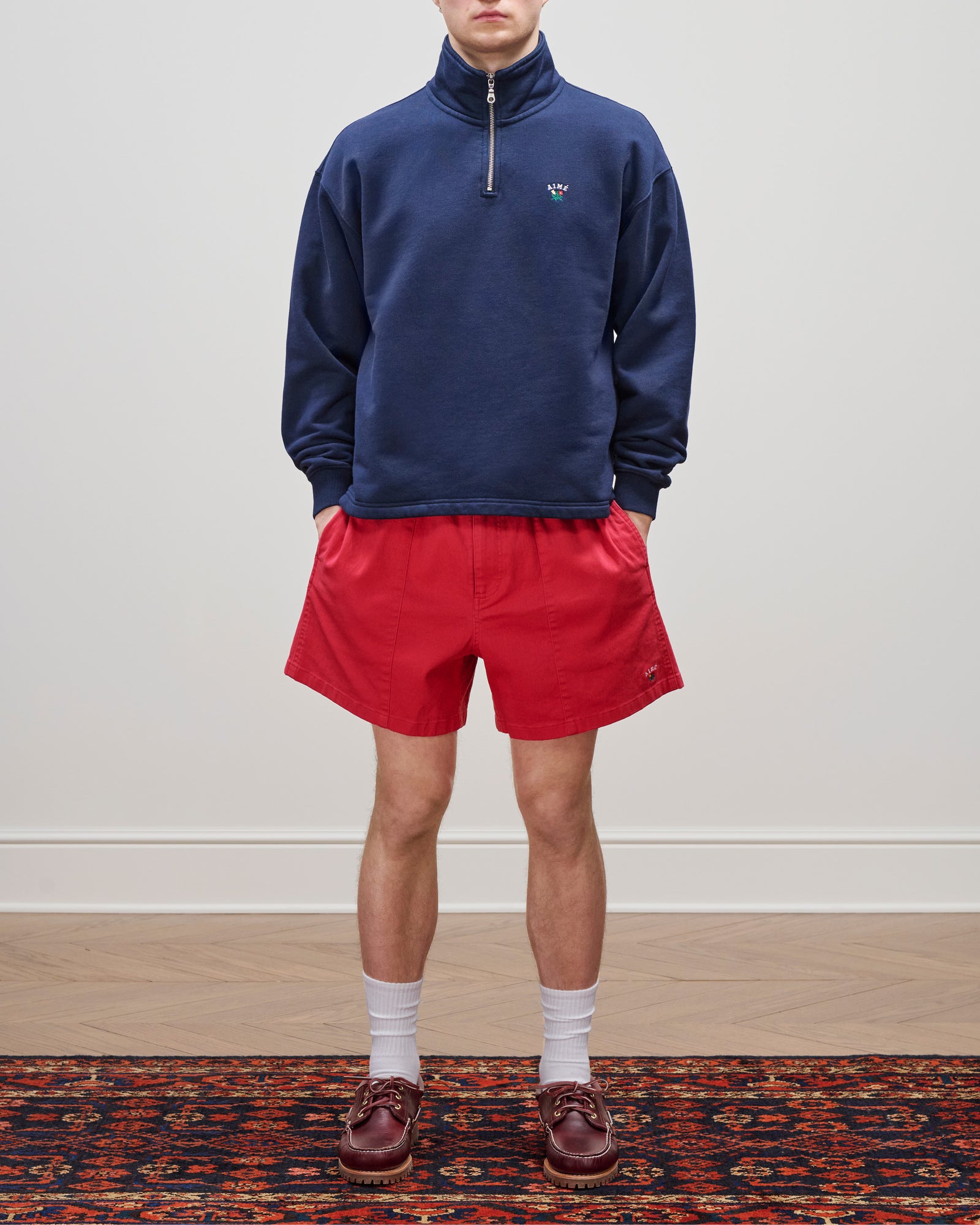Person wearing a navy Crest Quarter Zip Pullover, red shorts, white socks, and brown loafers on a patterned rug.