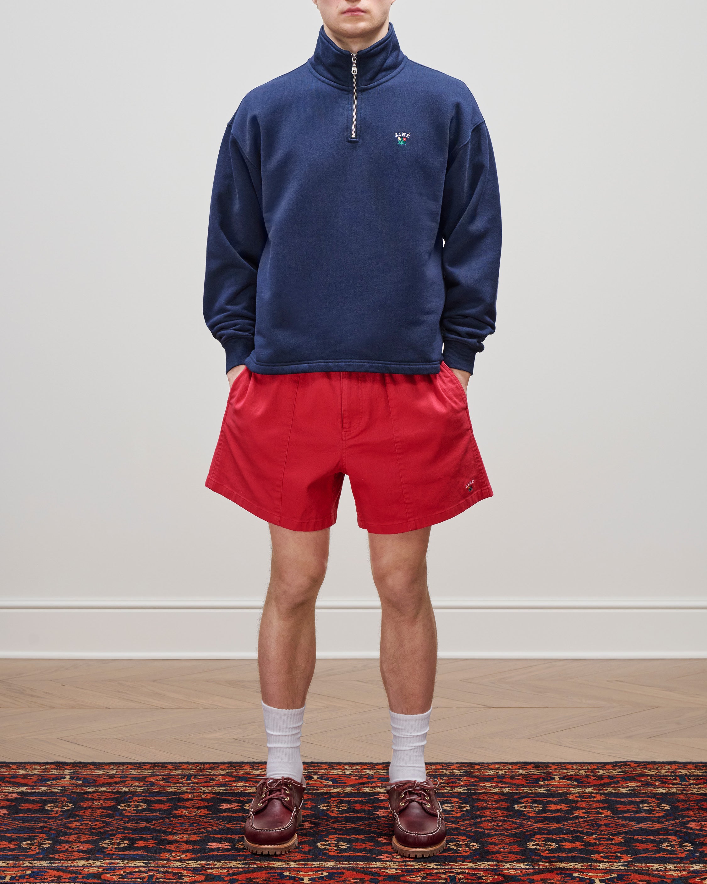 Person wearing a navy Crest Quarter Zip Pullover, red shorts, white socks, and brown loafers on a patterned rug.