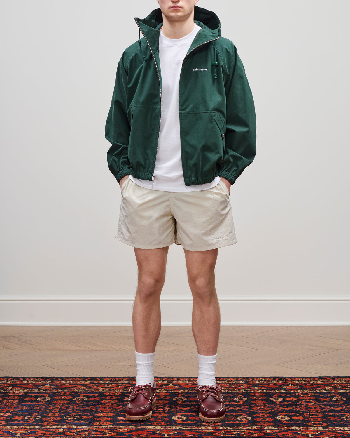 A person wears a green hooded jacket, white tee, Core Nylon Short in beige, white socks, and brown shoes on a rug.