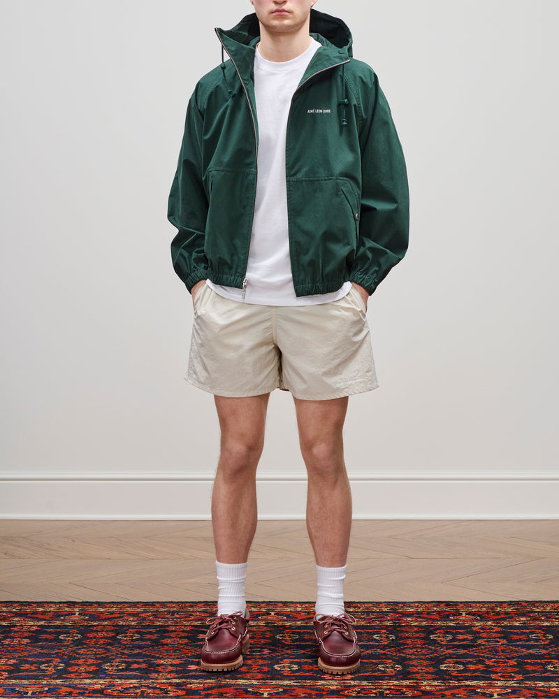 A person in a green Micro Logo Lightweight Jacket, white tee, beige shorts, and brown shoes stands on a patterned rug.