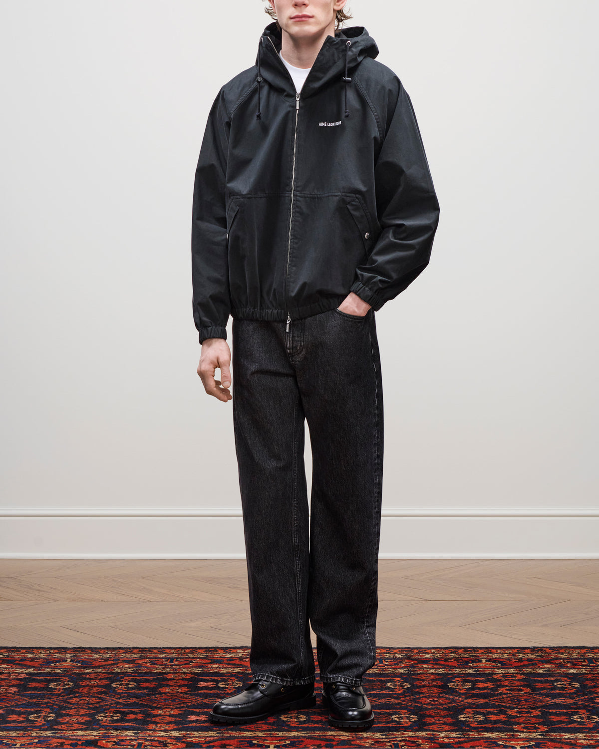 Person wearing a Micro Logo Lightweight Jacket, dark jeans, and black shoes stands on a patterned rug by a plain wall.
