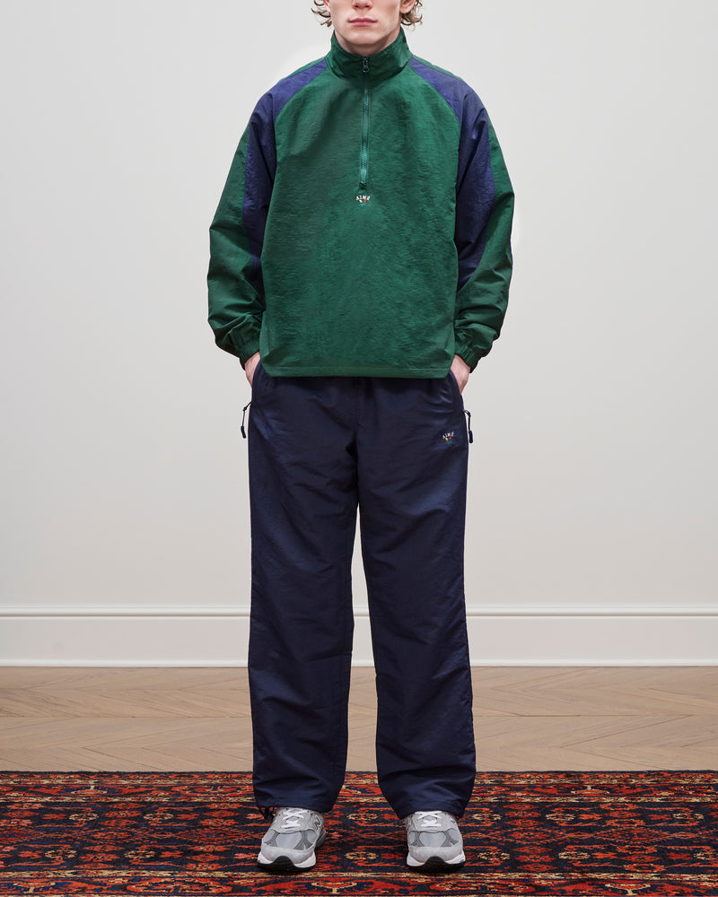 Person wearing the Crest Nylon Half-Zip Pullover in green and navy with gray sneakers, standing on a patterned rug.