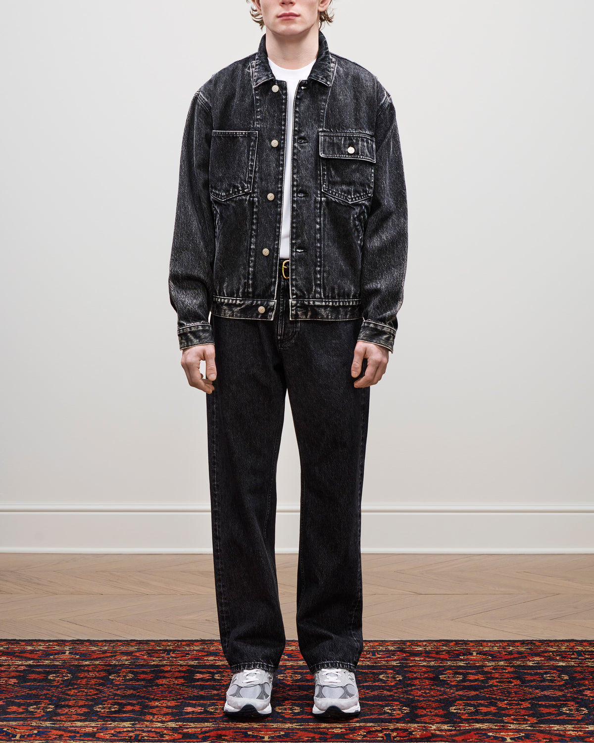 Person in a black Denim Trucker Jacket, pants, white tee, and gray sneakers stands on a patterned rug.