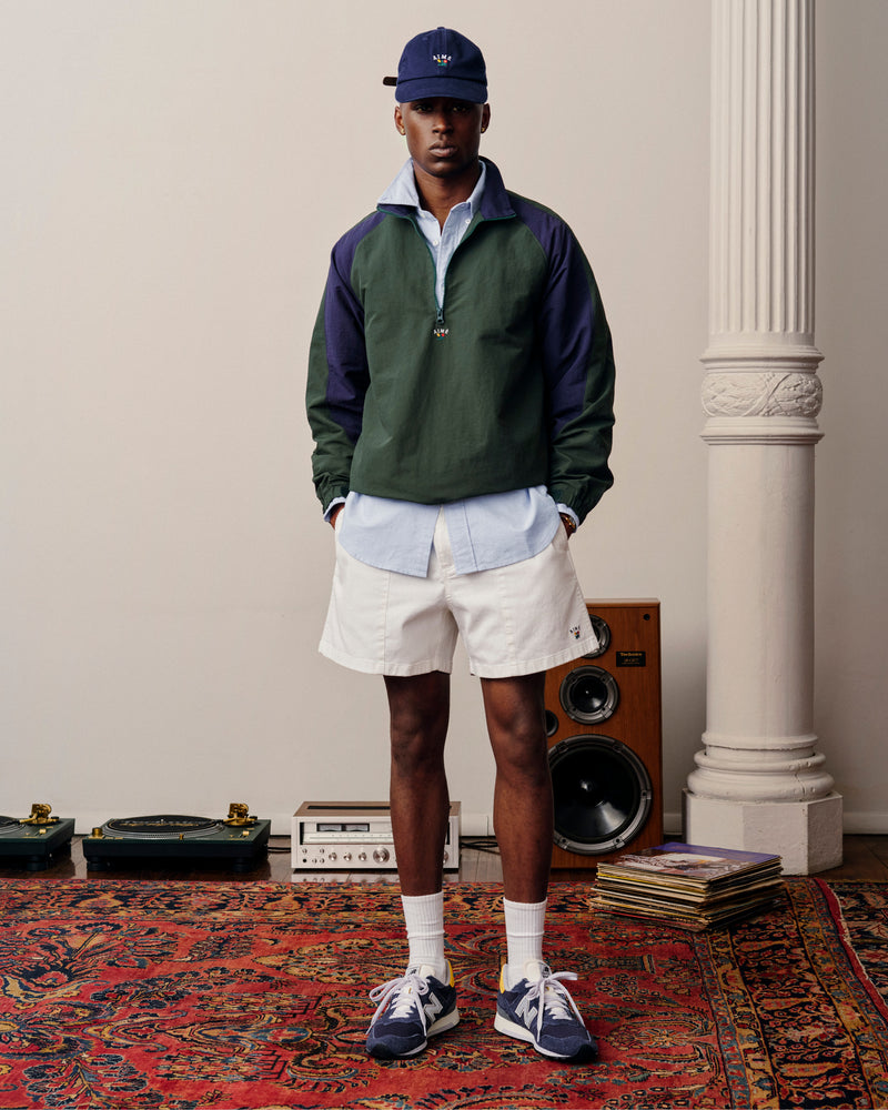 A man in a Crest Nylon Half-Zip Pullover, blue shirt, white shorts, and sneakers stands on a patterned rug by speakers.
