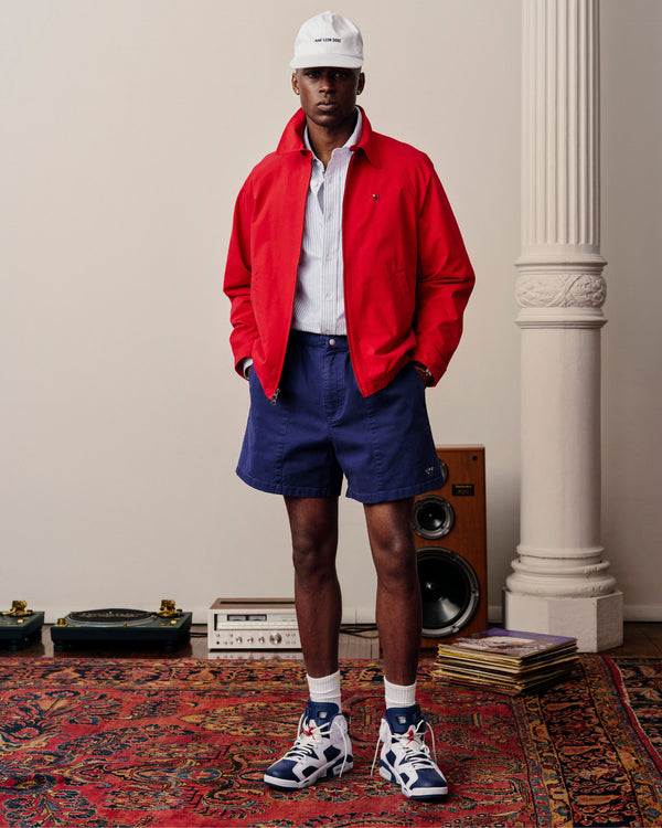 A man in Crest Casual Shorts, a red jacket, and sneakers stands indoors on a patterned rug near records and speakers.