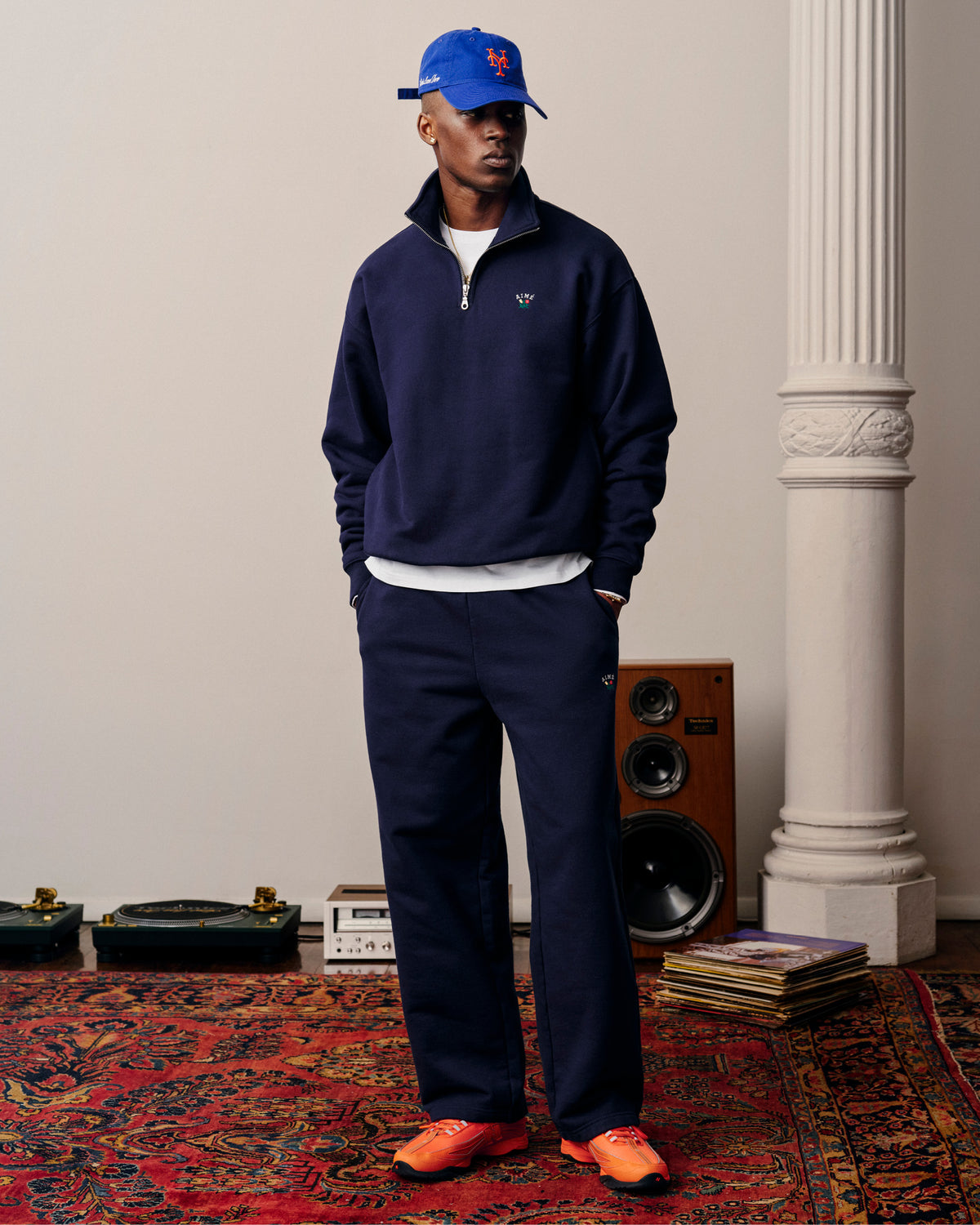 A man in Crest Sweatpants, a blue cap, and orange sneakers stands on a patterned rug near speakers and vinyl records.