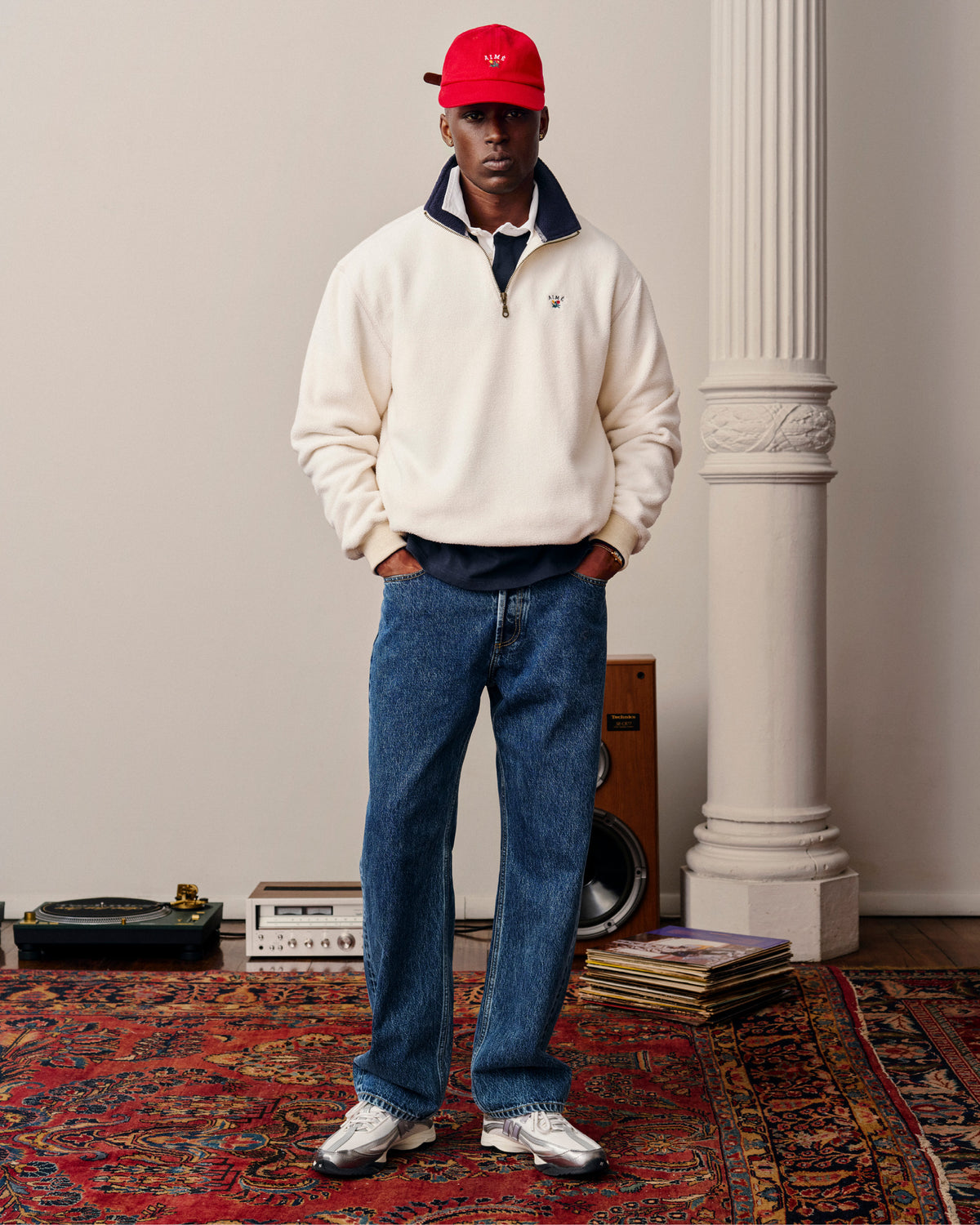 A man in a Crest Quarter-Zip Fleece Pullover, blue jeans, and red cap stands on a patterned rug near audio equipment.