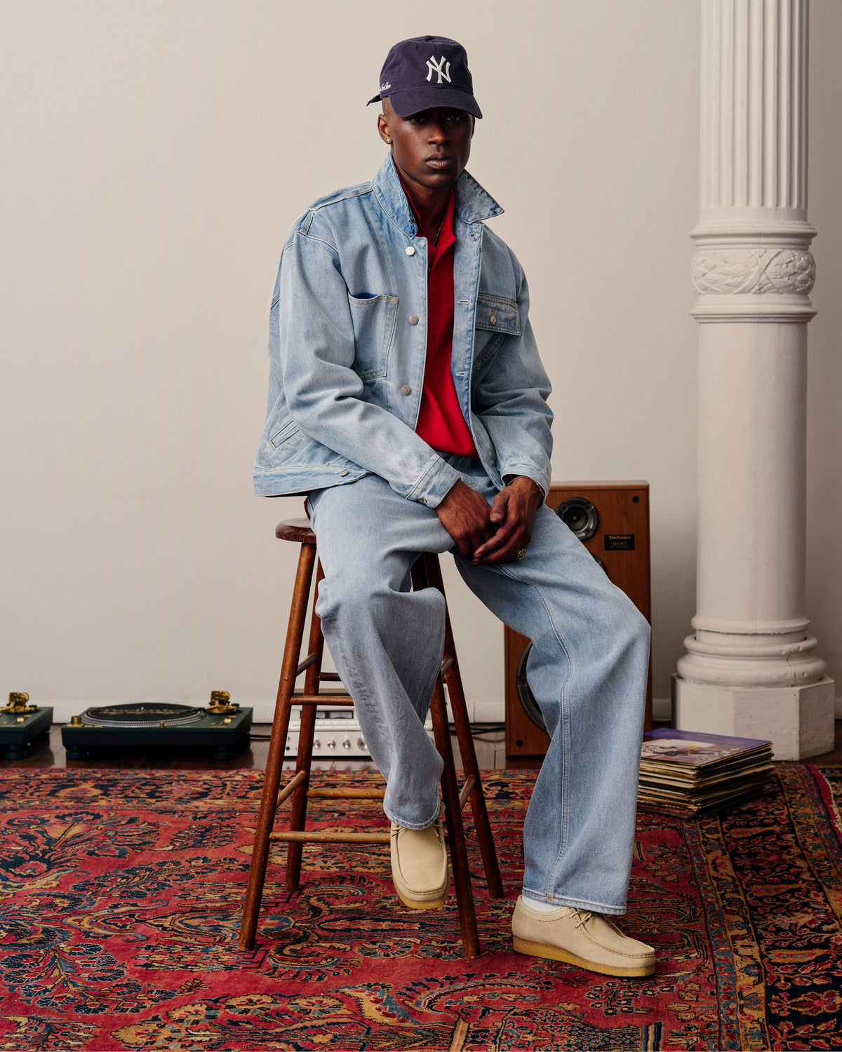 A person in a Long-Sleeve Crest Piqué Polo, light denim jacket and jeans, and NY cap sits on a stool in a stylish room.