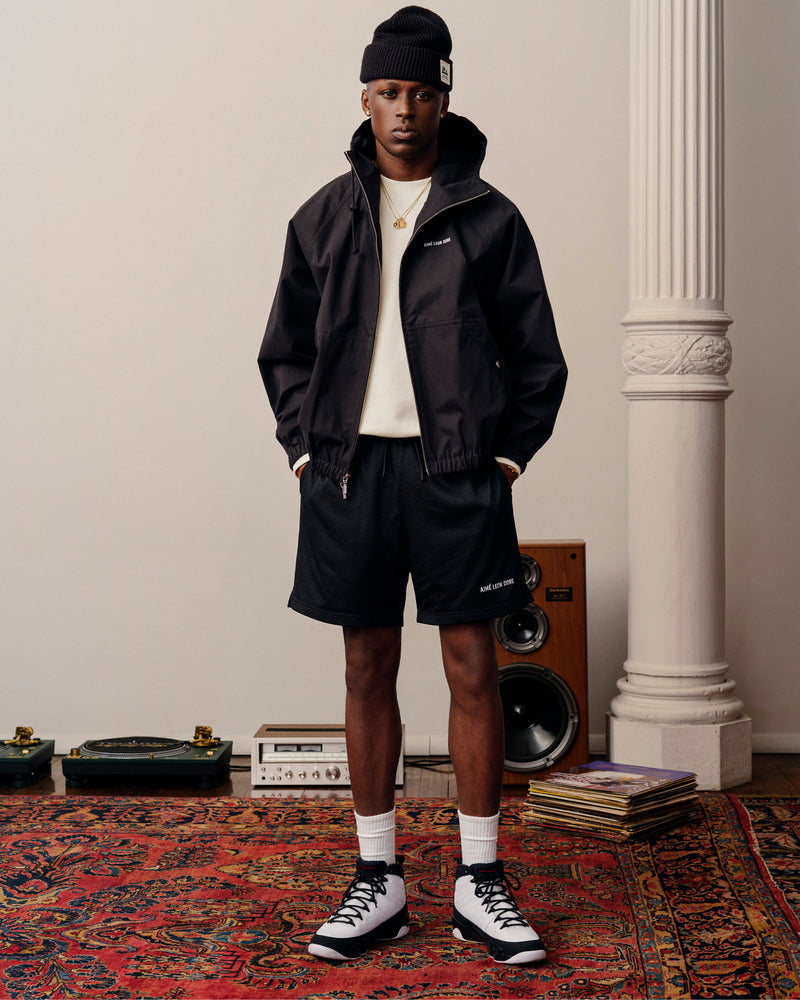 A man in a Micro Logo Lightweight Jacket, black shorts, and beanie stands on a patterned rug near vinyl records and a speaker.