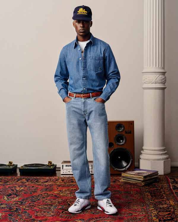 A man in a Crest Chambray Shirt, light jeans, sneakers, and navy cap stands on a red patterned rug by speakers and records.