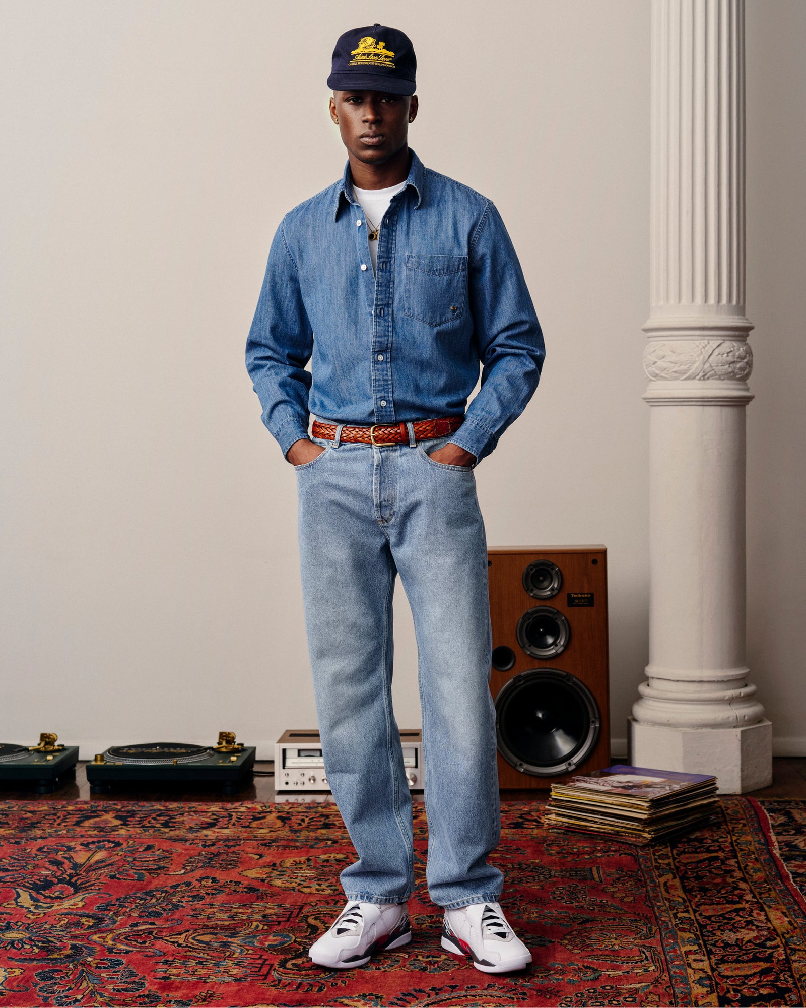 Man in ALD Straight Fit Denim, white sneakers, and cap stands on a patterned rug with records and speakers behind him.