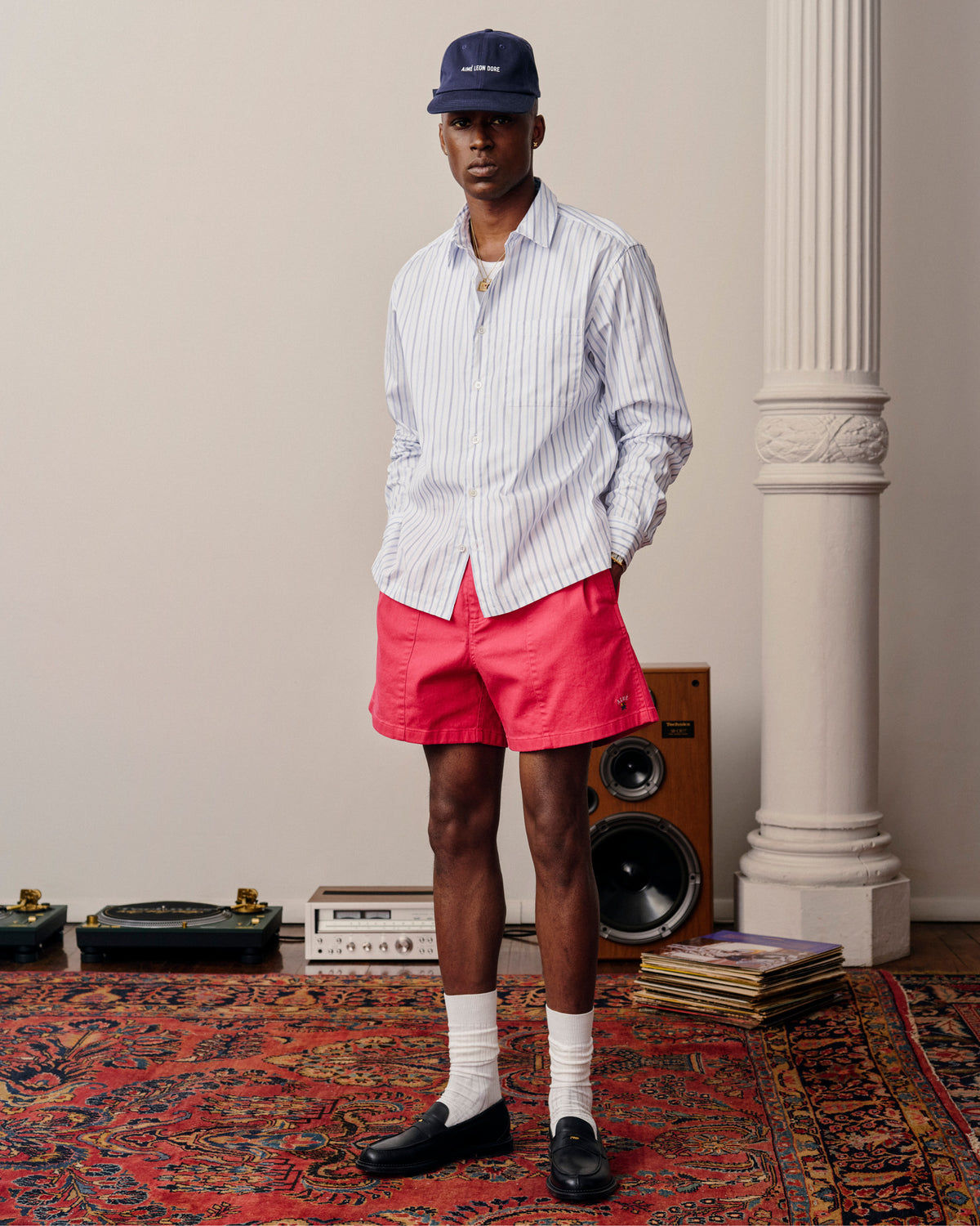 Man in a Poplin Casual Shirt, red shorts, cap, and loafers stands on a patterned rug near speakers and records.