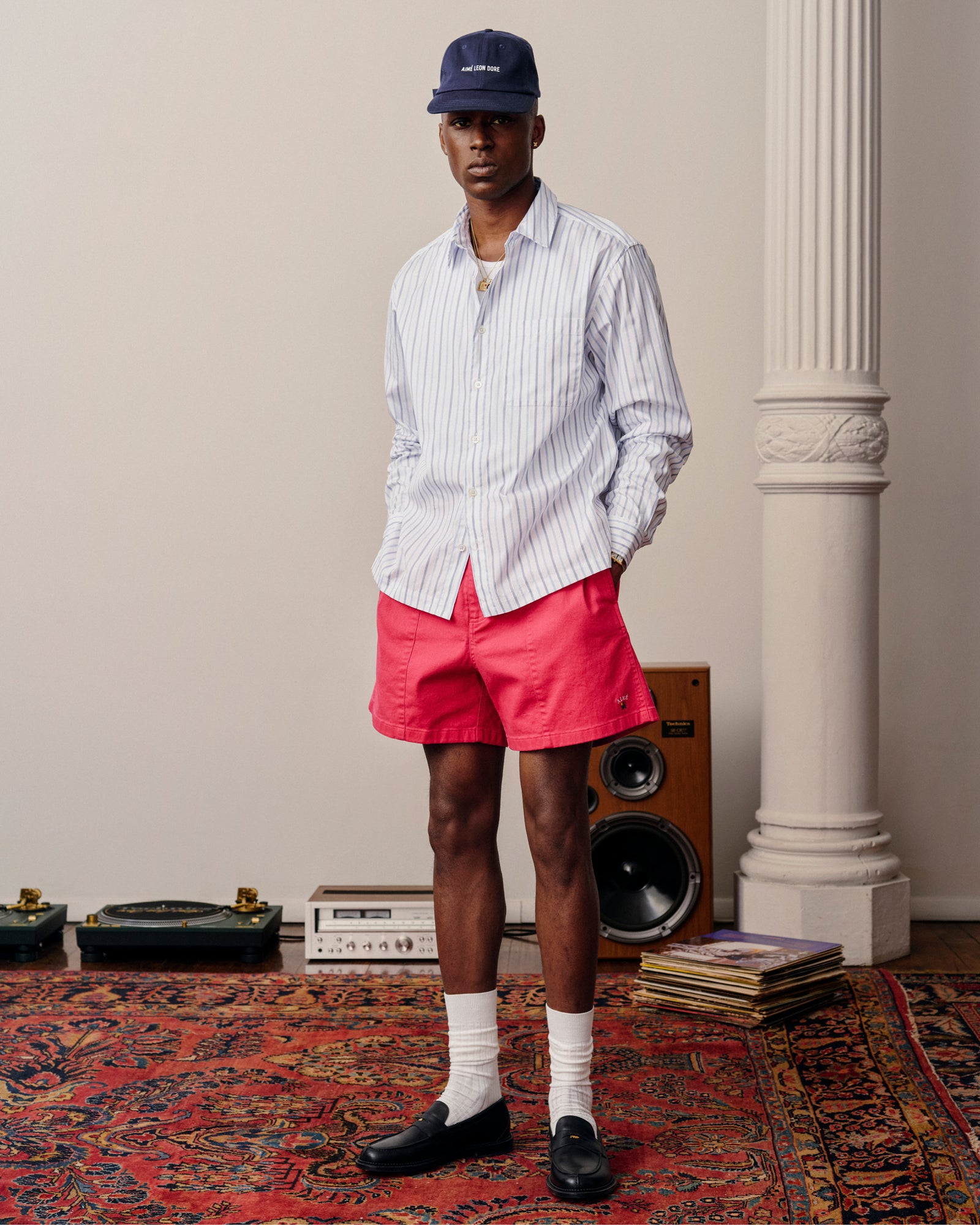 A man in a striped shirt, Crest Casual Short, and a cap stands on a patterned rug near speakers and vinyl records.