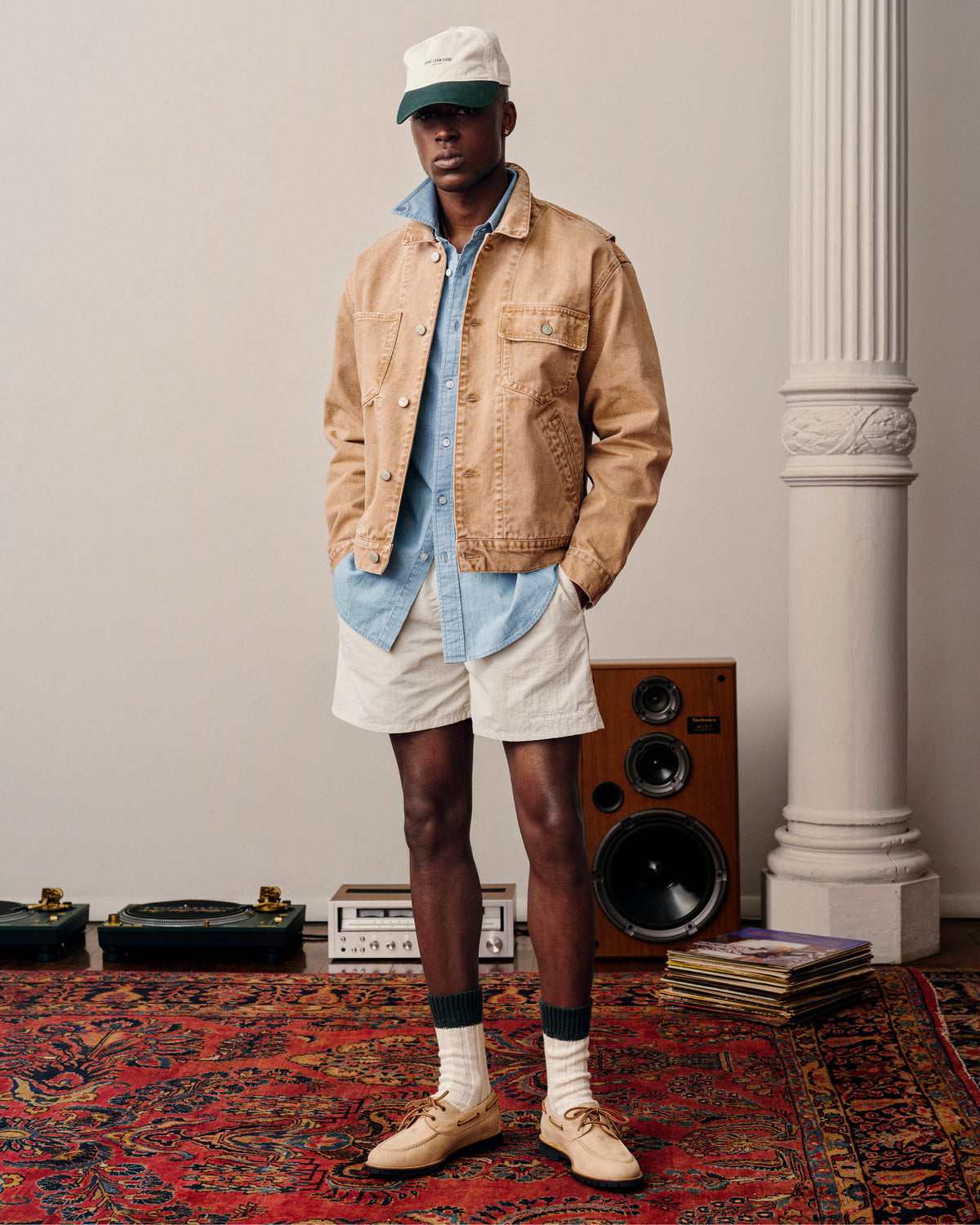A man in a tan jacket, shorts, Crest Chambray Shirt, and cap stands indoors on a patterned rug near speakers and records.