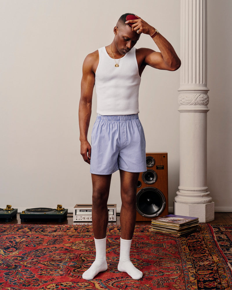 Man in a white tank top and Boxer Short 2-Pack stands on a patterned rug near speakers and vinyl records.
