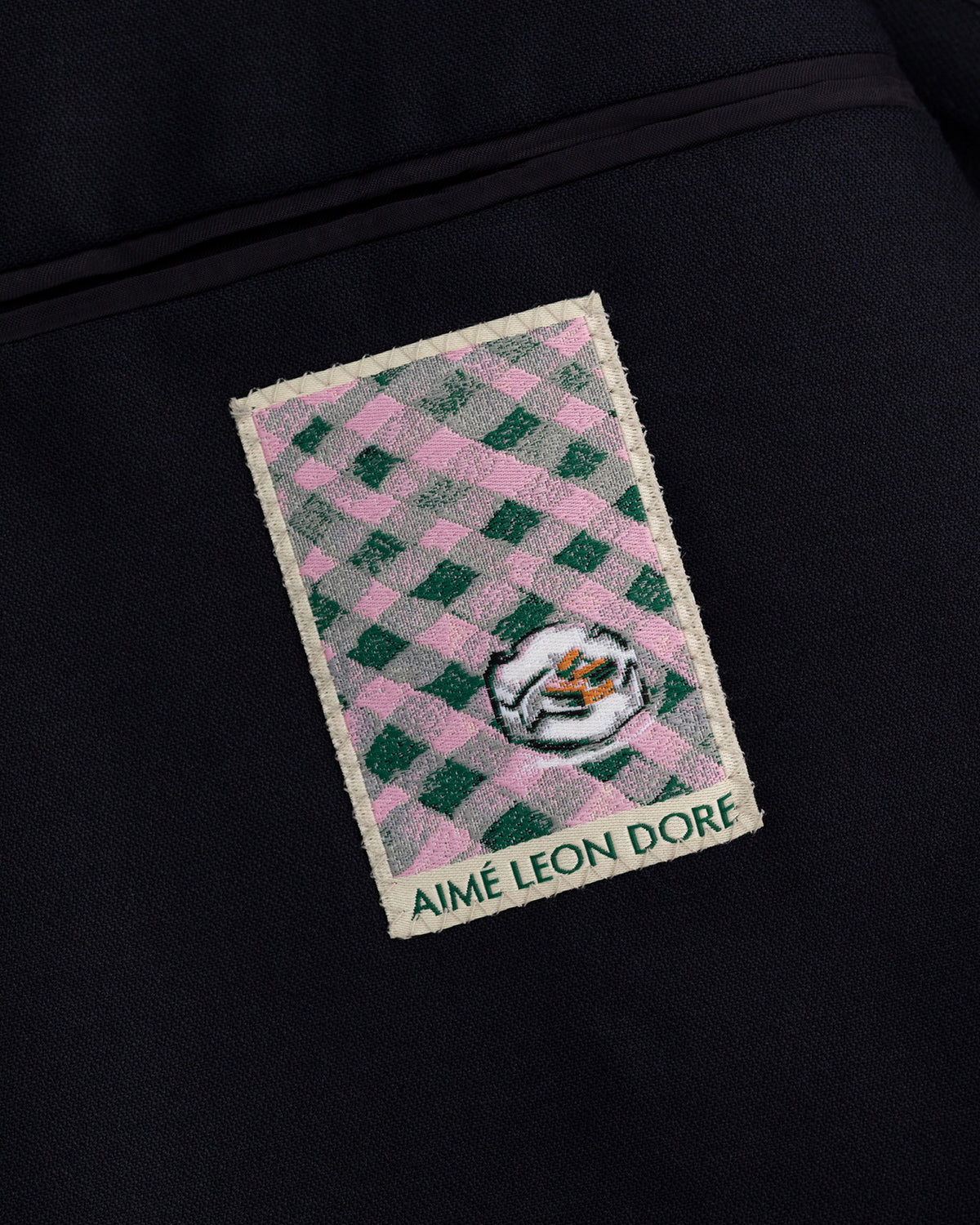 Rectangular Aimé Leon Dore patch with green-pink checks, white face, on dark Tropical Wool Suit Jacket.